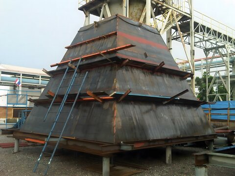 A large, pyramid-shaped metal structure with a weathered surface and orange support beams stands on a metal platform, surrounded by industrial equipment. A ladder leans against one side for access.