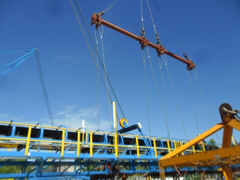 A large red beam is suspended by steel cables above a yellow conveyor belt with blue railings, set against a clear sky. Cranes and industrial equipment appear blurred in the background.