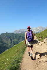 woman hiker walking hard with purple backpack on her shoulders and on Alpine mountains trail during summer season in northern Italy