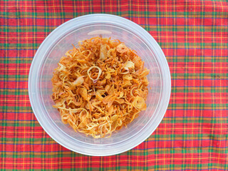 Deep-fried savory crispy sliced shallots, stored in a transparent plastic container, placed on a vibrant plaid red and green tablecloth, creating a colorful and traditional food presentation