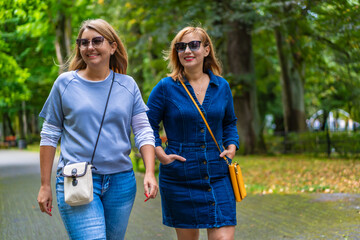 Portrait of two young beautiful smiling women dressed casual walking in city park on sunny day, trees in background. Meeting of friends. Walk in park.