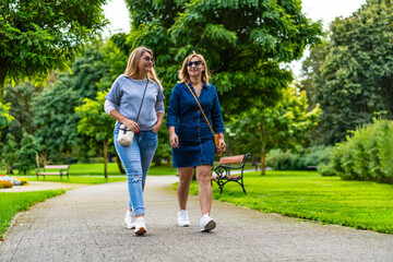 Two young beautiful smiling women dressed casual walking in city park on sunny day, trees in background. Meeting of friends. Walk in park.