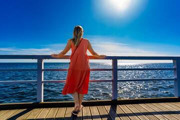 Summer vacation. Woman wearing flowing orange dress standing on wooden pier in seaside resort on...