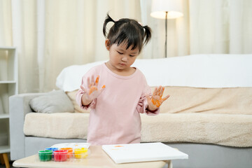Naklejka premium Asian toddler girl hands covered in orange paint, exploring the sensation and colors during finger painting. various paint colors, expressing creativity in indoor environment, learning art and play.