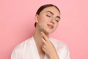 Beautiful young woman doing facial massage with roller on pink background