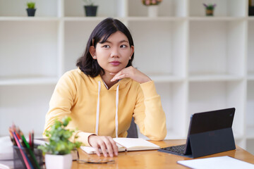 A young woman in a yellow hoodie is sitting at a desk, looking thoughtful while working on her...