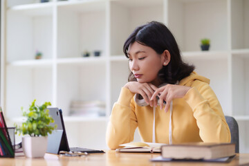 Obraz premium A young woman in a yellow hoodie is sitting at a desk, working on her laptop and holding a pen. She appears to be thinking, with plants nearby and a bright, modern workspace around her