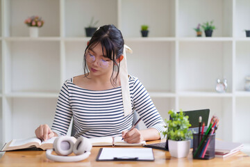 A young woman wearing glasses and a striped shirt is sitting at a desk, writing in a notebook. She appears focused, with an organized indoor setting around her and shelves in the background