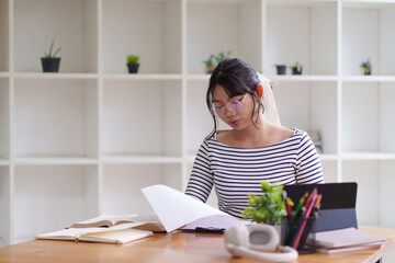 A young woman in a striped shirt is sitting at a desk, looking at a piece of paper while working on her laptop. She is in a bright room with large windows, surrounded by natural light