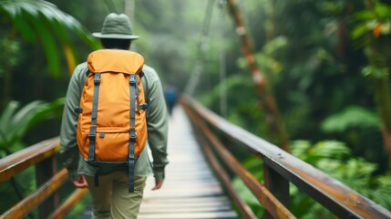 A hiker wearing a green jacket and an orange backpack crosses a wooden bridge in a dense tropical forest, surrounded by lush greenery.