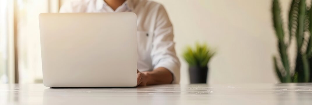 Business professional in a white shirt works on a laptop in a well-lit office with natural sunlight and minimalistic decor, including a small plant in the background.
