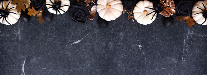 Halloween top border of black flowers, gold leaves and white pumpkins with spiders. Overhead view over a dark black stone banner background. Copy space.