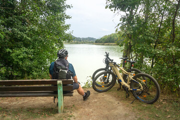 Älterer Mann sitzt auf einer Holzbank am See mit Helm auf dem Kopf und zwei Fahrrädern an seiner...