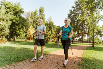 Happy senior couple nordic walking in park wearing sportswear, together exercising outdoors