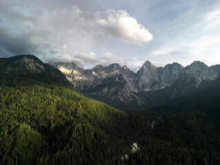 Fototapeta premium mountain village nestled at the base of towering, rugged mountains covered in dense forests. Sunlight breaks through the clouds, casting beautiful shadows over the vibrant green fields and small, scat
