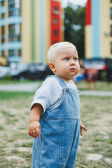 A 1-year-old baby boy in denim overalls is walking on the street. Children's oversized overalls. Stylish little boy on the playground.