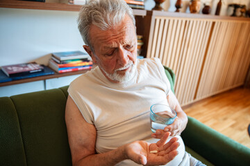 Elderly man holding pills and a glass of water sitting on a couch at home