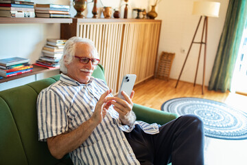 Senior man relaxing on couch and using smartphone at home
