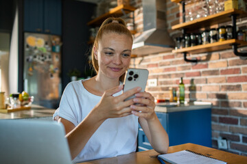 Young woman using smartphone in kitchen while working from home