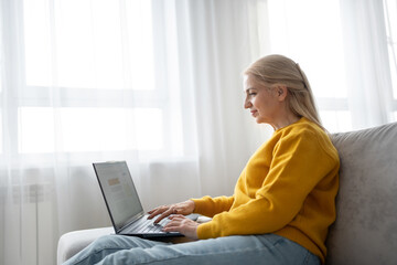 Naklejka premium Blonde woman in sweater types on laptop in welllit living room, creating productive workspace