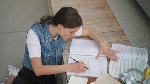Video of focused beautiful student in front of university building or campus, studying outdoors, reading textbook, preparing for final exam.