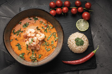 Tom Yam Kung Spicy Thai soup with shrimp in a bowl on a dark background and red pepper, cilantro rice on a black mica board. top view