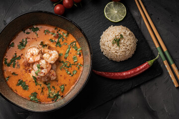 Tom Yum Kung Spicy Thai soup with shrimp in a bowl on a dark background and red pepper, rice and lime with cilantro and chopsticks on a black mica board. top view