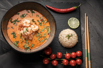 Tom Yum Kung Spicy Thai soup with shrimp in a bowl on a dark background and red pepper, rice and lime with cilantro and chopsticks on a black mica board. top view
