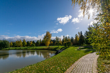 A park with a pond and a picturesque landscape on a sunny autumn day. Paved paths and a green lawn are strewn with fallen leaves.