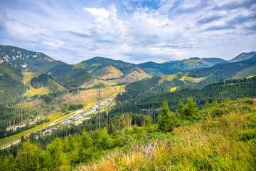 A panoramic view captures the beauty of the Jasna Mountain Resort nestled under Chopok Mountain in the Low Tatras. Lush greenery and mountain slopes create a picturesque landscape on a bright day.