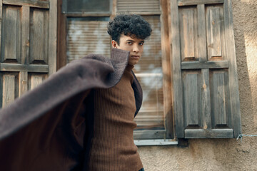 Fashion young man in brown sweater standing in front of old building with open and wooden shuttered...