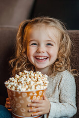Naklejka premium A Caucasian preschooler girl, full of joy, holding a bucket of popcorn while watching a comedy movie in a cozy cinema. Her giggles are infectious.