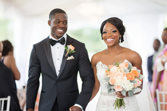 A cheerful Black bride and groom walking down the aisle after their wedding ceremony, holding hands and smiling brightly at the camera.