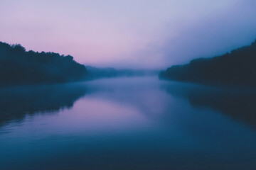 A serene, foggy river scene with a gradient sky shifting from soft lavender at the horizon to a deep, moody blue above. The river is partially obscured by mist, with its surface reflecting the gradien