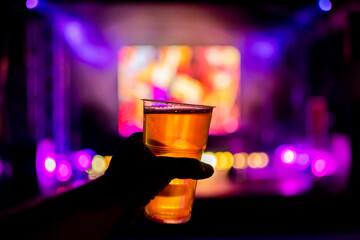 A close-up of a hand holding a glass of beer, with colorful bokeh lights and a blurred television screen in the background. Perfect for themes of leisure, celebration, and nightlife.