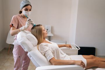 Relaxed pretty blonde female client having hair light therapy with modern innovation apparatus. Trichologist in mask and gloves performing hair treatment procedure with spectrum phototherapy device.