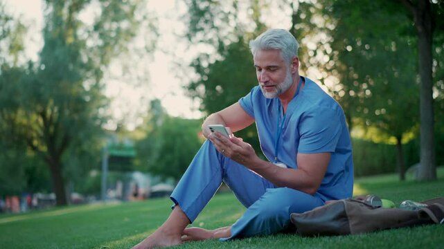 Handsome doctor in scrubs sitting on grass, relaxing with phone after long day in work. Job satisfaction, feeling contentment with his job. Mental health of healthcare worker.
