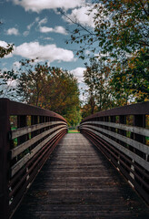 Old Wooden Bridge Crossing a Calm River in a Lush Nature Park, harming Wooden Bridge Overlooking a River in a Serene Nature, Sunny Summer Day with Trees and Clouds