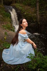Fototapeta premium A beautiful young girl with long hair and a blue dress near a forest waterfall