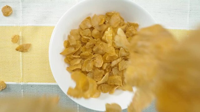 Close up of a healthy yellow cereal dropping in to bowl. Top down aerial view of cornflake falling into white bowl at white and black background. Macrography. Slow motion. Breakfast concept. Pabulum.