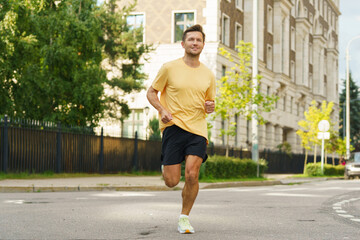 A man in a yellow shirt and black shorts is jogging along a quiet street with trees and buildings.