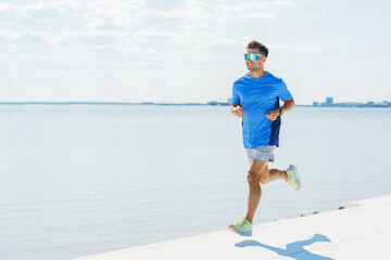 A man runs along a waterfront in bright sunlight, wearing sportswear and reflective sunglasses.
