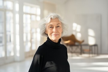 Fototapeta premium Smiling elderly woman in black turtleneck stands in a bright minimalistic room with stylish furniture, enjoying a moment of joy
