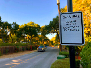 License plates monitored by camera warning sign is displayed on a quiet residential street with a passing vehicle. Selective focus. Close up.
