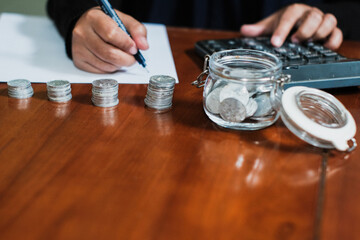 Saving concept with pile of the coins and coins on the jar glasses and a hand holding pen taking note the amount of money with calculator in the wooden table, Savings and investing concept.