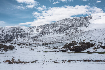 Cold snow-white landscape of snowy valley with alpine lake against snow-covered rocky mountain range under clouds in blue sky. Glacial lake, pure white snow and stones in high mountains in sunny day.