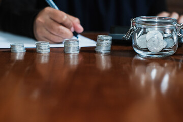 Saving concept with pile of the coins and coins on the jar glasses and a hand holding pen taking note the amount of money with calculator in the wooden table, Savings and investing concept.