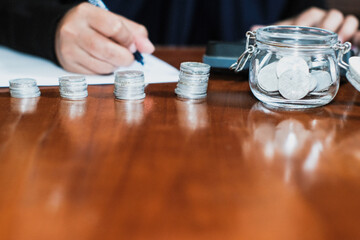 Saving concept with pile of the coins and coins on the jar glasses and a hand holding pen taking note the amount of money with calculator in the wooden table, Savings and investing concept.