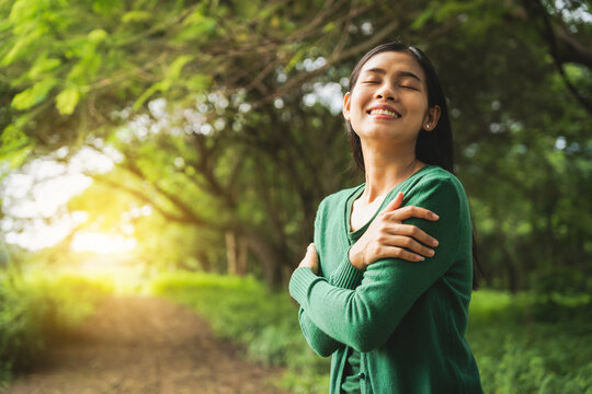 confident asian woman embraces self-love, standing peacefully in serene park at sunset. joyful smile, she enjoys fresh air, breathing deeply, radiates calm and positivity in tranquil surroundings.
