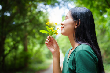 A cheerful young asian woman enjoys a sunny day outdoors, holding a vibrant bunch of yellow flowers. Smiling in a green field, she embodies the joy and freedom of summer in the countryside.